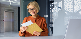 A woman smiles as she pulls a paycheck out of an envelope while sitting at a work desk.