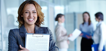 In the foreground, a smiling woman holds a clipboard.  In the background, her employees discuss work.
